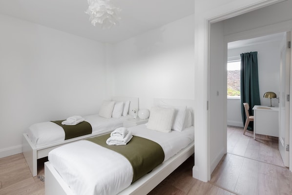 A modern hotel room with two single beds, a desk, a chair, and a view of the outdoors through a window with curtains. - Brandon House 3 bedroom house VRBO (Dingle)