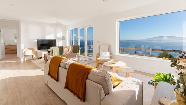 Una sala de estar moderna con un gran ventanal con vista al mar, un sofá con una manta y un televisor de pantalla plana.