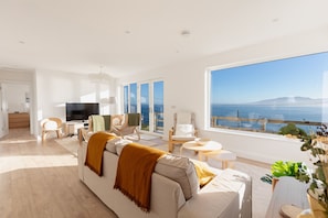 A modern living room with a large window overlooking the sea, a sofa with a throw blanket, and a flat-screen TV. - Brandon House 3 bedroom house VRBO (Dingle)