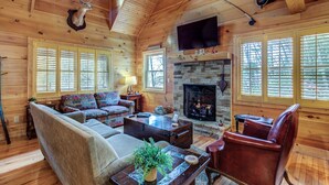 A cozy log cabin interior with a fireplace, wooden walls, a sofa, a leather chair, and a mounted deer head. - Blowing Rock Cabin | Hot Tub, Fire Pit, Fall Views (Blowing Rock)