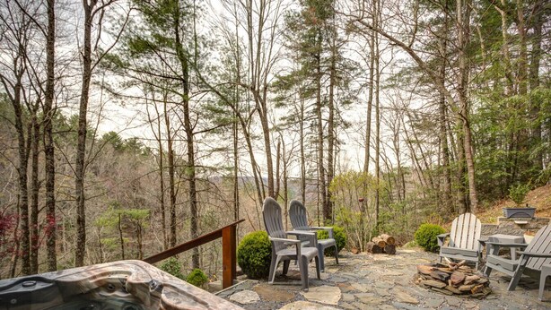 A hot tub surrounded by Adirondack chairs and a stone deck in a forested area. - Blowing Rock Cabin | Hot Tub, Fire Pit, Fall Views (Blowing Rock)