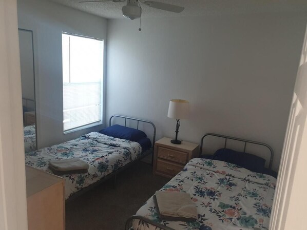 A small, simple room with two single beds, a nightstand with a lamp, and a window providing natural light.