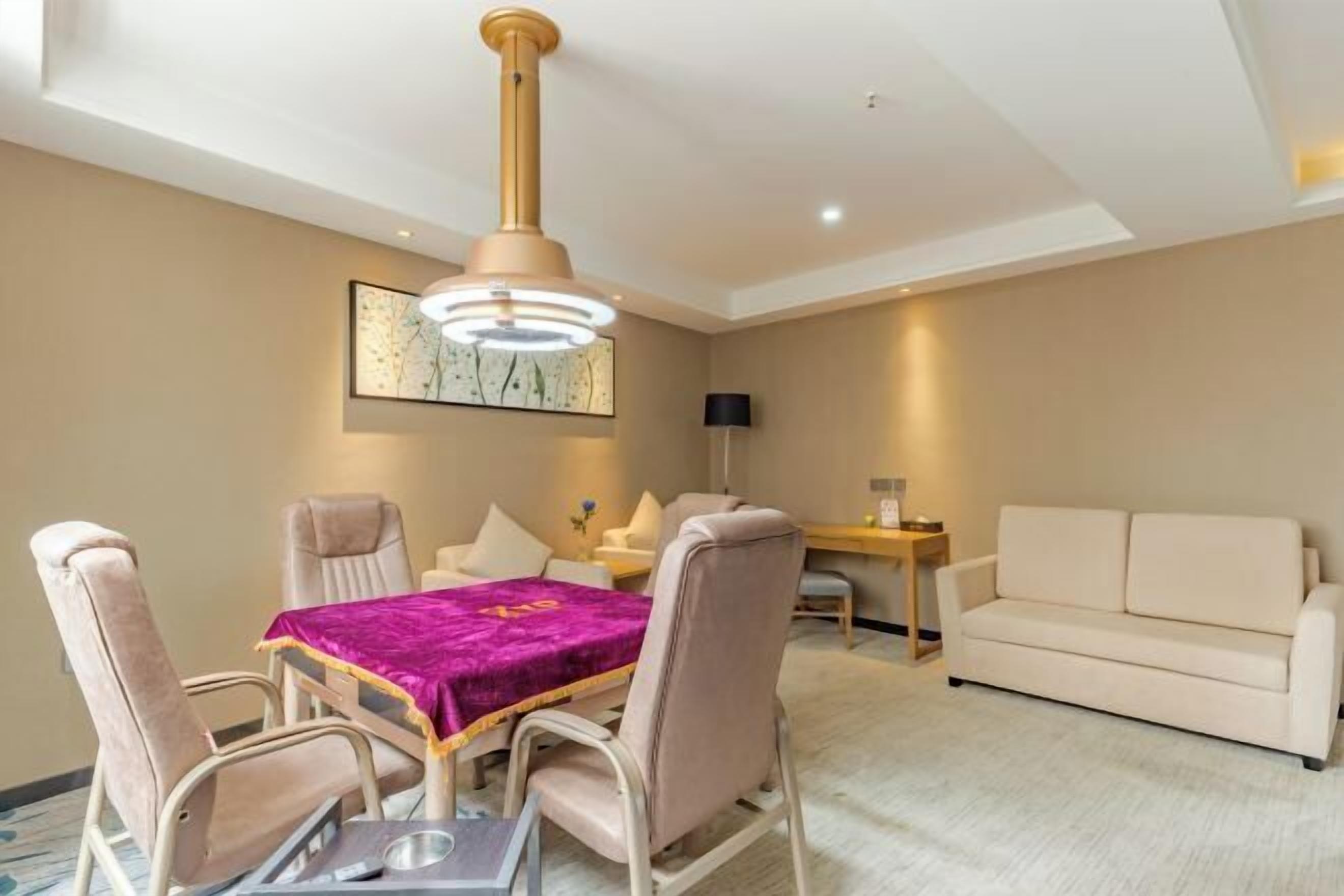 A hotel room with a dining area featuring a table with a purple tablecloth, beige chairs, and a ceiling light fixture.