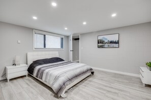 Une chambre moderne comprenant un lit, des tables de chevet, une fenêtre avec des stores, un tableau accroché au mur et une porte donnant sur une autre pièce.