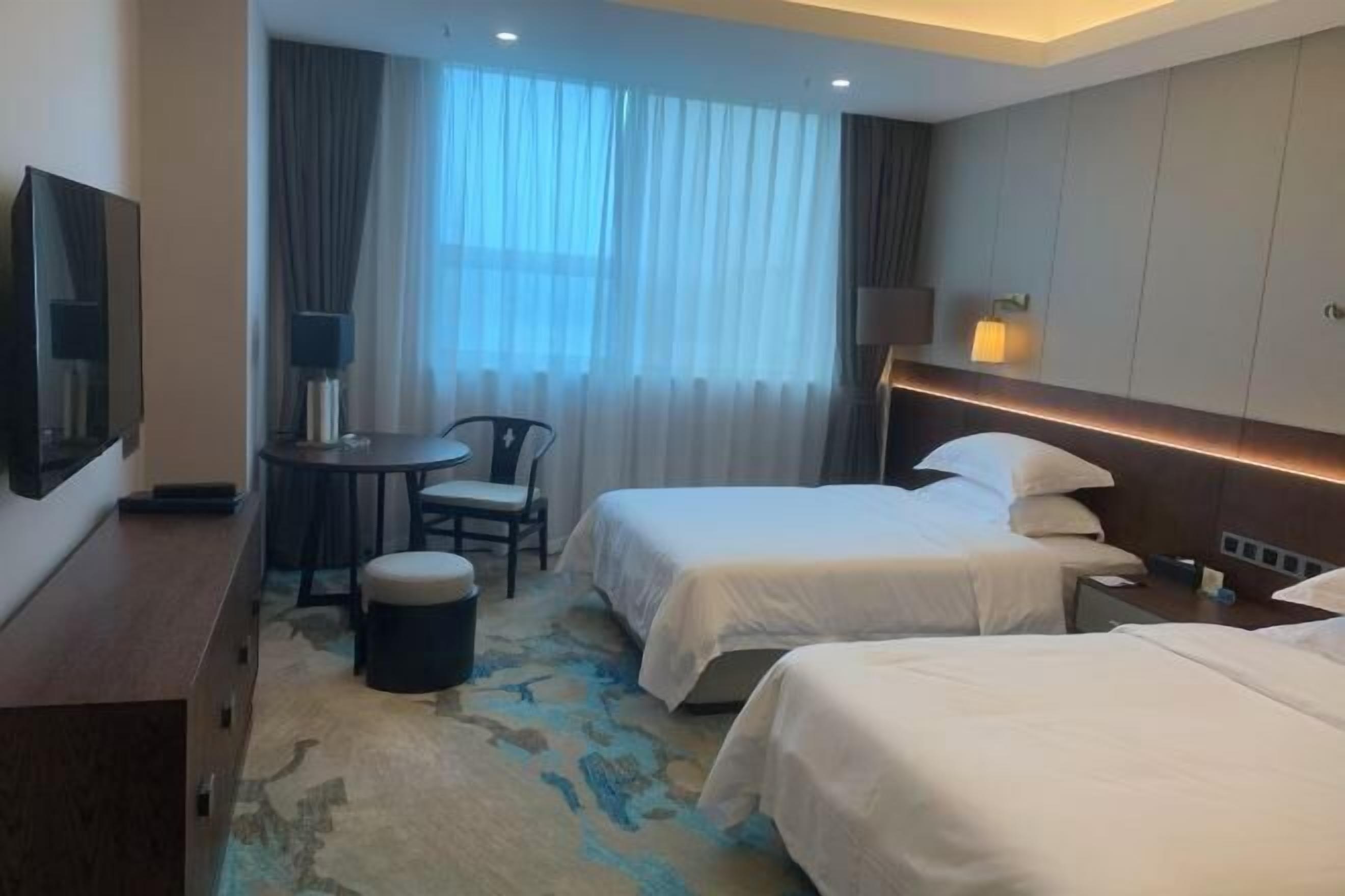 A hotel room with two beds, a desk with a chair, a TV, and a large window with curtains.
