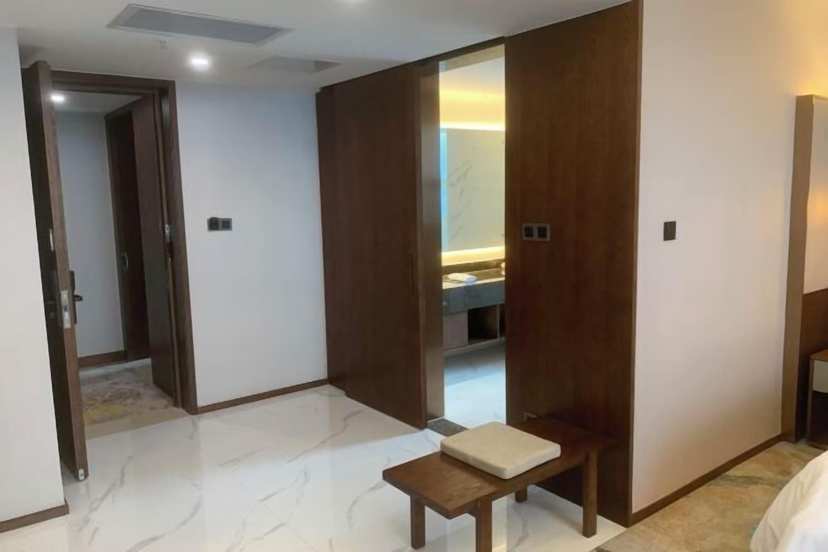 Quarto de hotel com guarda-roupa de madeira, um banco, uma mesinha e um banheiro visível com espelho.