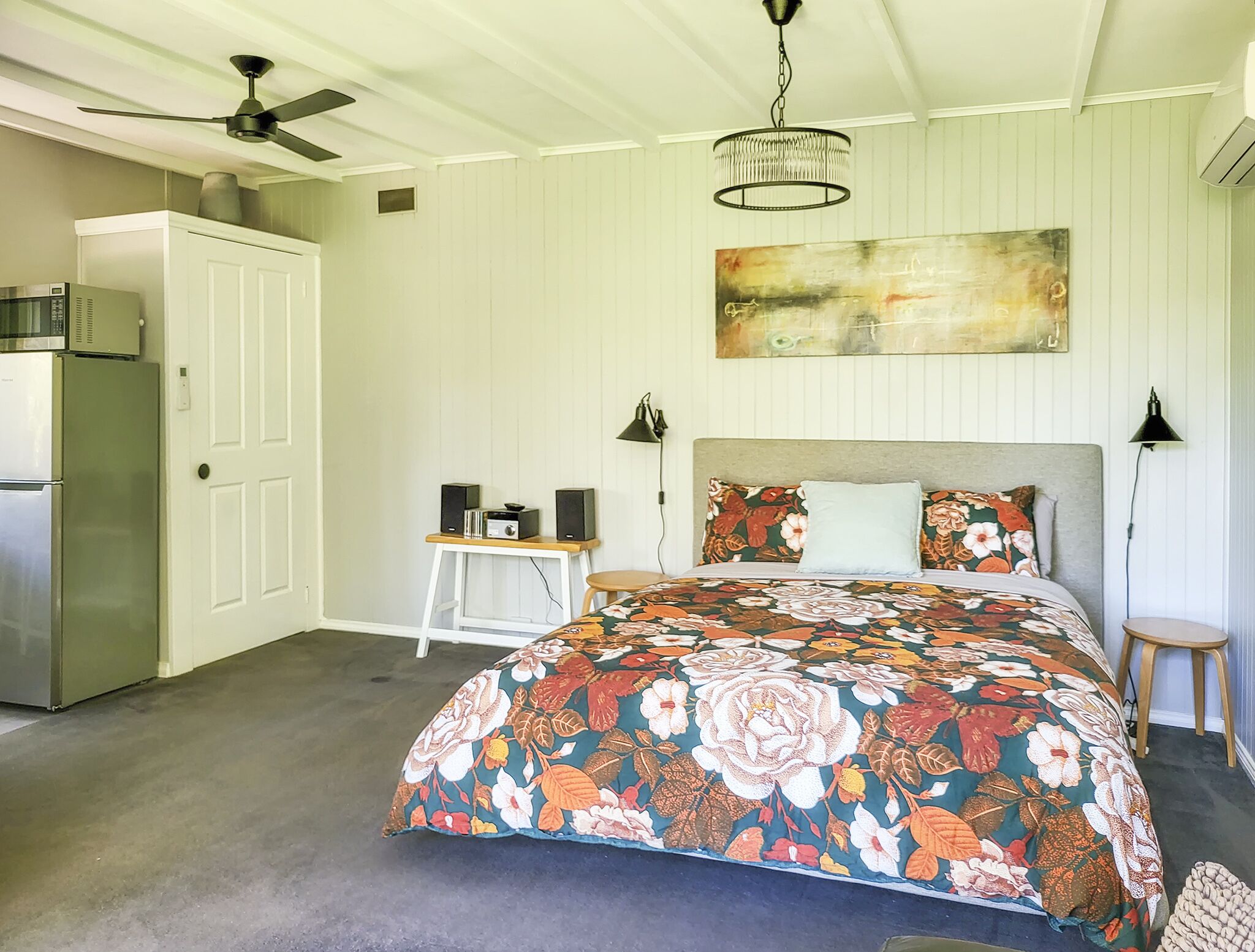 Ein Schlafzimmer mit einem großen floralen Bettüberwurf, einem Deckenventilator, einer Wandklimaanlage, einem kleinen Tisch mit Lautsprechern und einer Wandlampe.