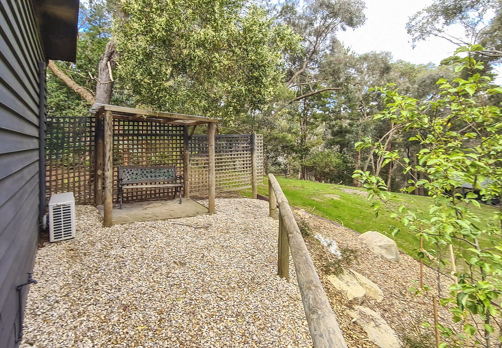 Uma área de estar coberta ao ar livre, com um banco, cercada por uma vedação de madeira e chão de cascalho, em um jardim com árvores e gramado.