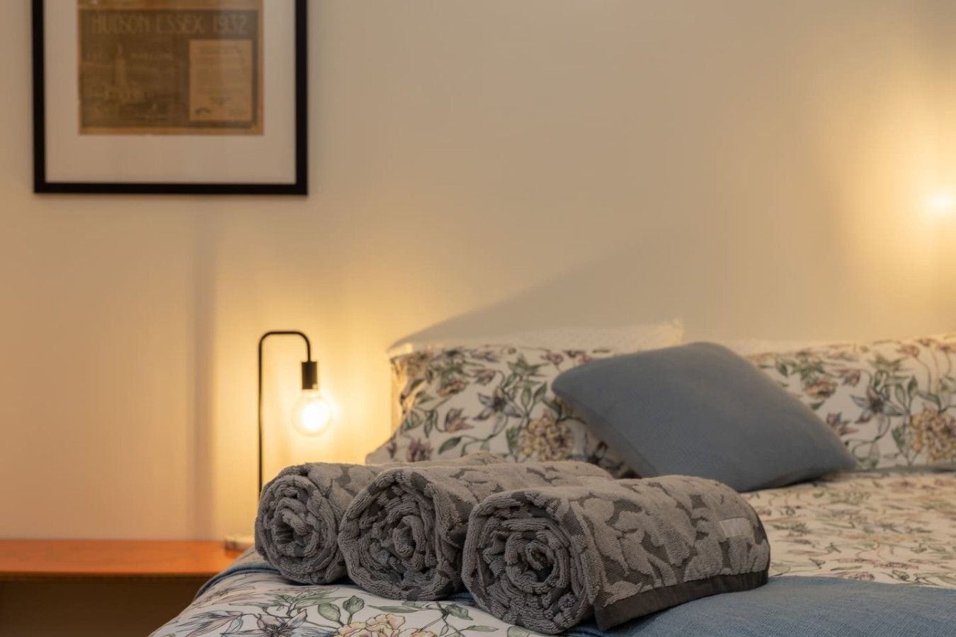 Una cama bien tendida con toallas enrolladas, una almohada y una lámpara fijada a la pared.