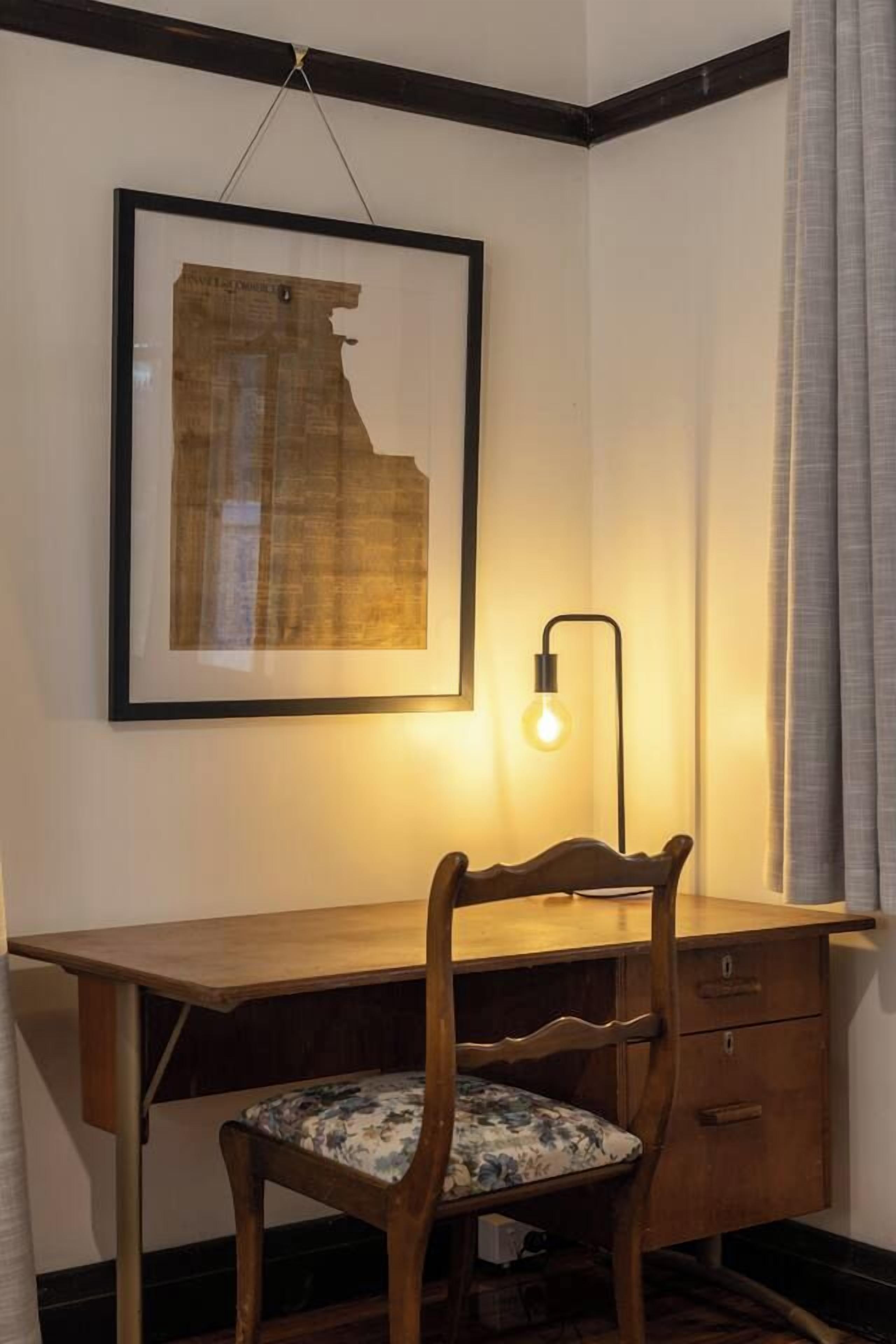 A corner room with a desk, chair, framed map, and a wall-mounted lamp.
