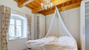 A bedroom with a bed, wooden ceiling beams, a window with curtains, and a chandelier. - Vacation home Frilo by Interhome (Broumov)