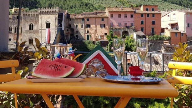 Une table avec une pastèque, un drapeau et un plateau contenant des verres et une rose rouge, donnant sur un village aux bâtiments colorés et sur une colline en arrière-plan.