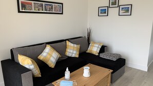 Een moderne woonkamer met een zwarte bank, geel- en grijze kussens, een houten salontafel met desinfecterende handgel en een mondkapje.