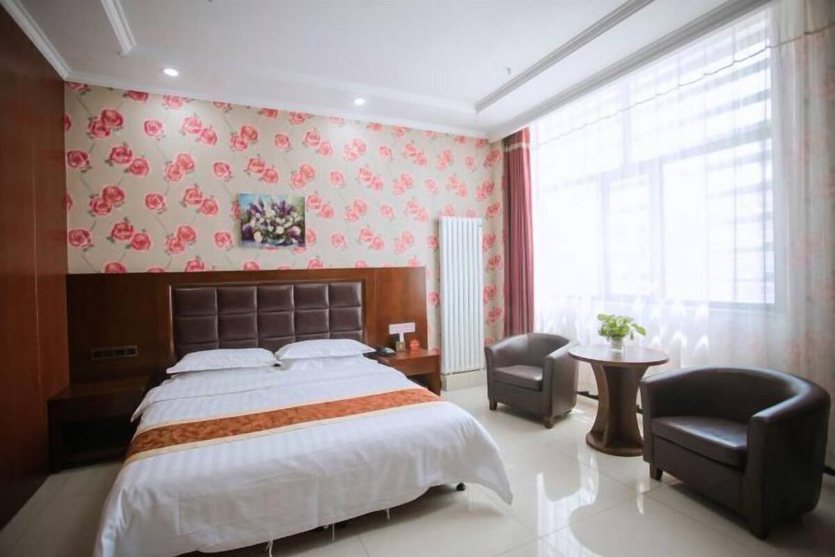 Habitación de hotel con una cama grande, cabecera, mesita de noche, escritorio, dos sillas y una ventana con cortinas.