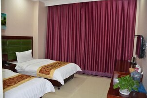 Habitación de hotel con dos camas, cabecero verde, ventana grande con cortina roja, escritorio de madera con una planta y un espejo.