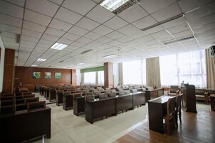 Un salón de conferencias con filas de mesas y sillas, ventanas amplias y un techo alto con luminarias a la vista.