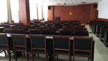 A conference room with rows of wooden chairs and tables, a projector screen, and a red emblem on the wall.