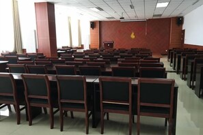 Un salón de conferencias con filas de sillas y mesas de madera, una pantalla de proyector y un emblema rojo en la pared.