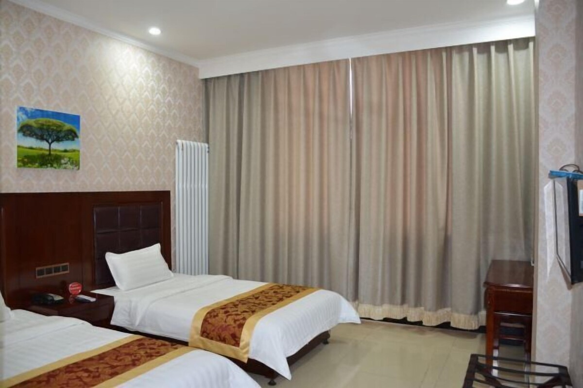 Habitación de hotel con dos camas, cabecera de madera, un cuadro de un árbol y una ventana grande con cortinas.