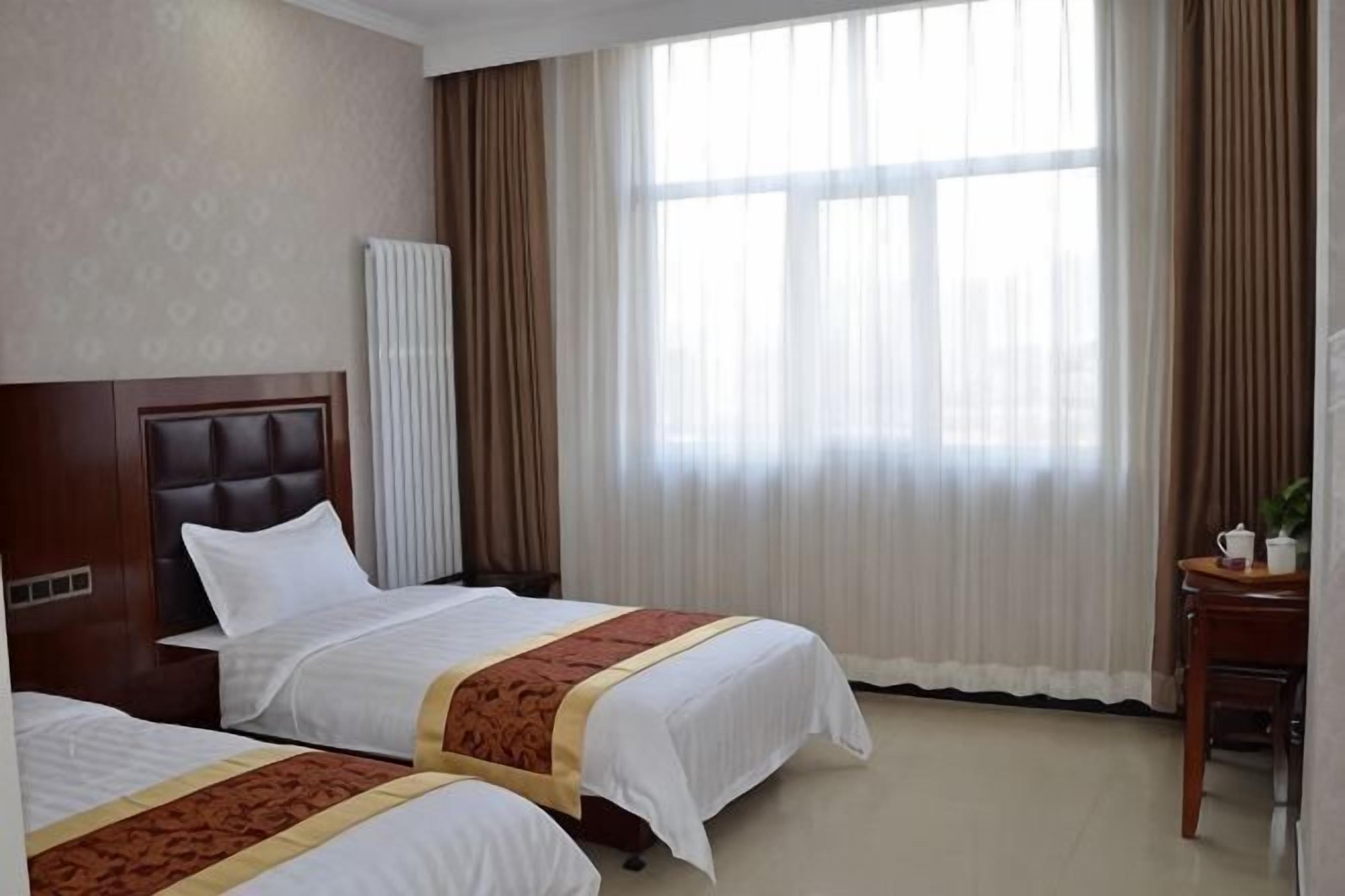 A hotel room with two beds, a wooden headboard, a window with curtains, and a small table with a plant.
