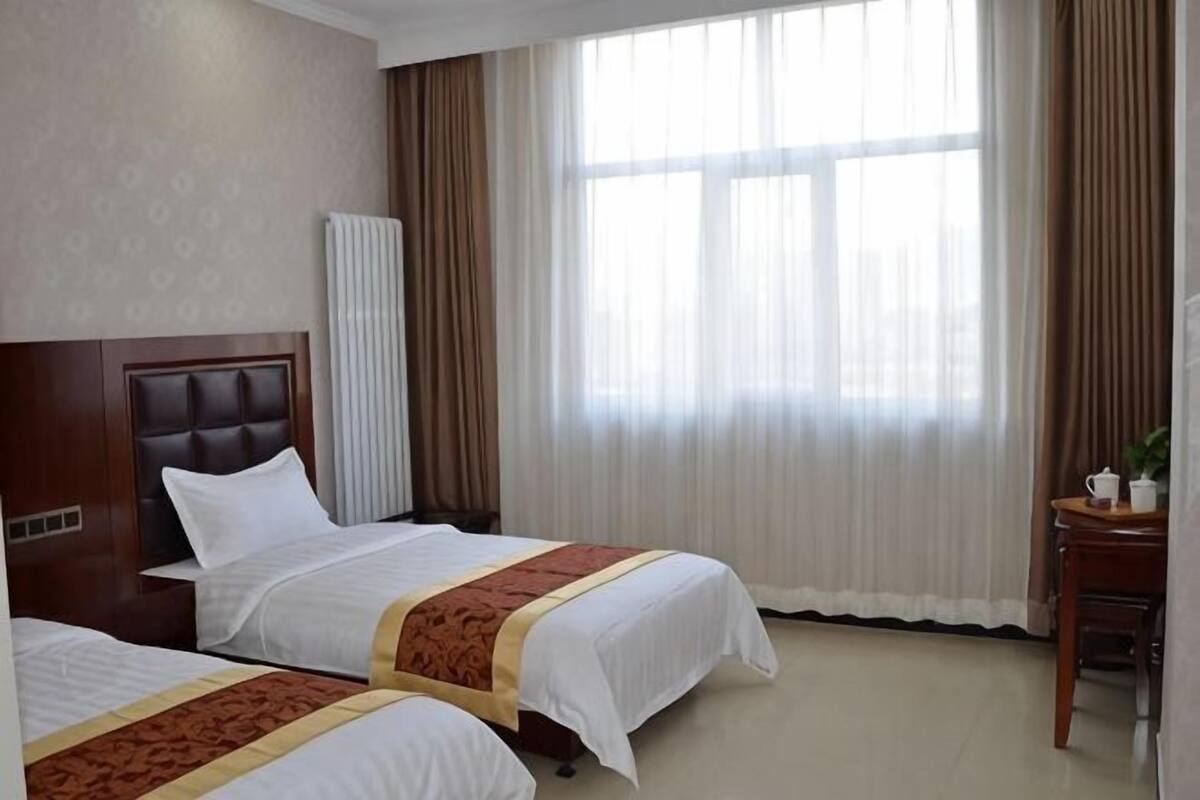 Habitación de hotel con dos camas, cabecera de madera, ventana con cortinas y una mesita con una planta.