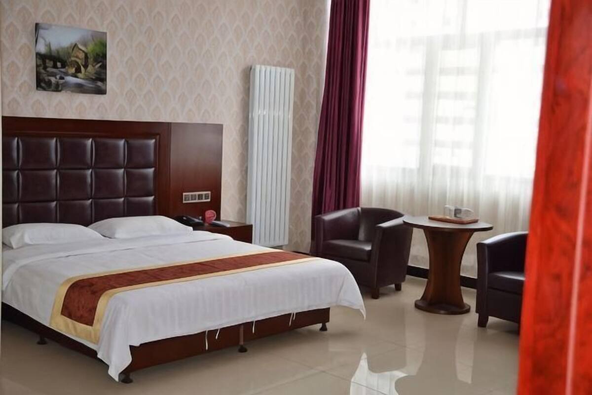 Habitación de hotel con una cama grande, una mesita de noche, una silla, una pequeña mesa y un ventanal con cortinas.