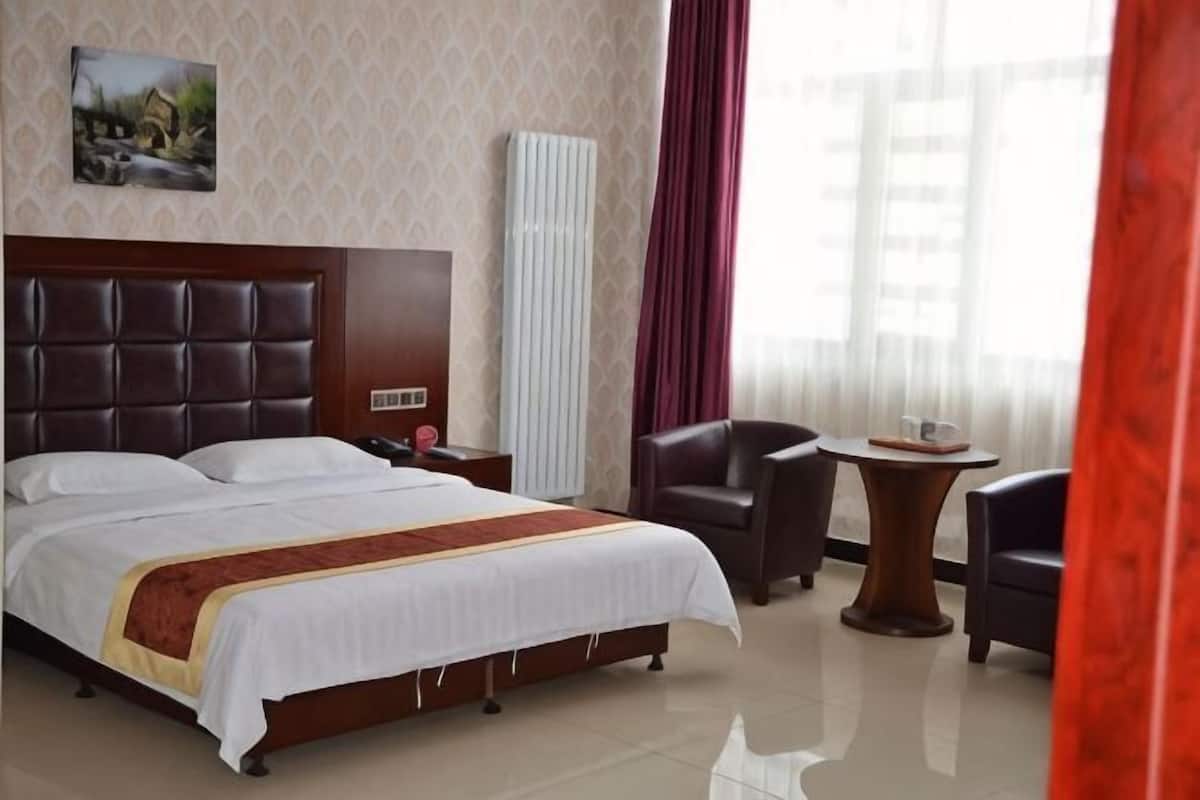 A hotel room with a large bed, a bedside table, a chair, a small table, and a window with curtains.