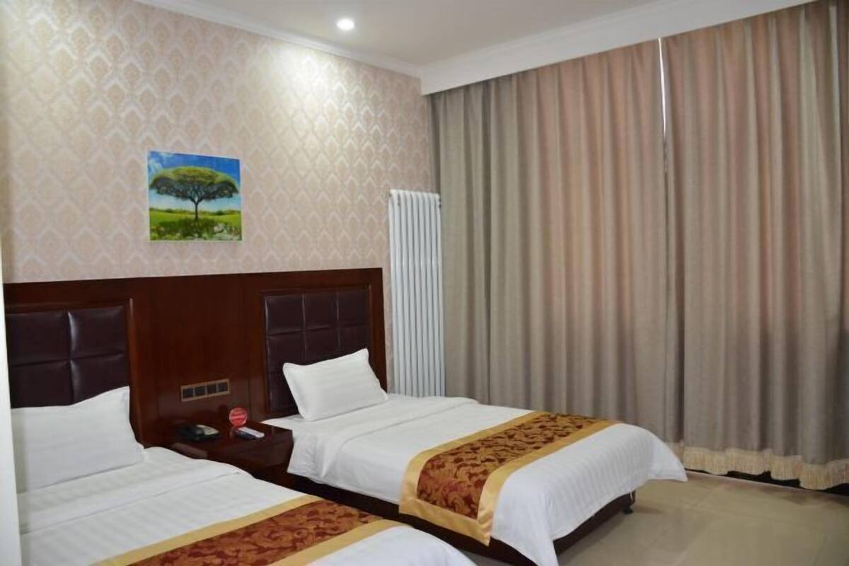 Habitación de hotel con dos camas, cabecero de madera, radiador y un ventanal con cortinas.