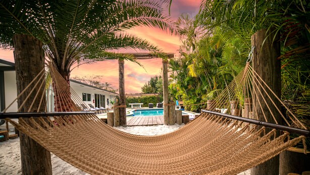 A hammock between wooden posts on a sandy beach with a pool and palm trees in the background. - MAGR LUXURY (Cutler Bay)