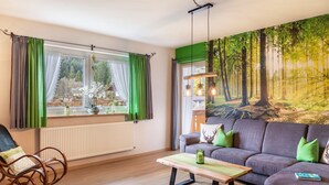 A living room with a sofa, a rocking chair, a table, and a large forest mural on the wall. - Holiday Apartment Waldruhe with Balcony, Mountain View & Wi-Fi (Garmisch-Partenkirchen)