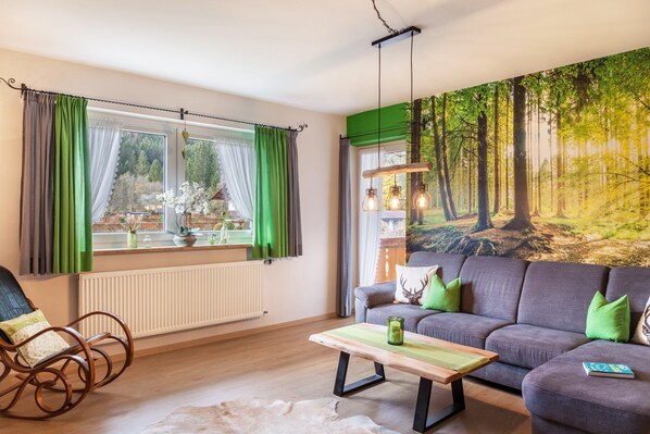 A living room with a sofa, a rocking chair, a table, and a large forest mural on the wall.