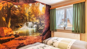 A room with a large mural of a forest waterfall, a bed with floral bedding, and a window with green curtains. - Holiday Apartment Waldruhe with Balcony, Mountain View & Wi-Fi (Garmisch-Partenkirchen)