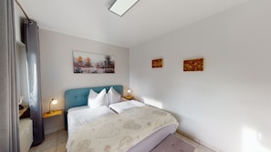 Une chambre à coucher comprenant un lit, deux tables de chevet, une commode, une fenêtre avec des rideaux et deux œuvres d’art accrochées au mur.