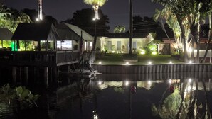 Ein Nachtbild eines Anwesens am Wasser mit einem Steg, einem Gebäude mit geneigtem Dach und einem gut beleuchteten Garten.