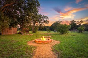 A circular fire pit in a grassy area with trees and a wooden structure in the background.