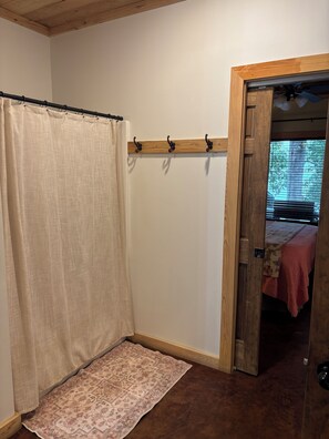 Une pièce avec un plafond en bois, un rideau beige, un porte-manteau en bois, un tapis à motifs et une porte donnant sur une autre pièce où se trouve un lit.