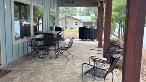 En overdækket terrasse med et bord og stole, en loftsvifte og et hus i baggrunden.