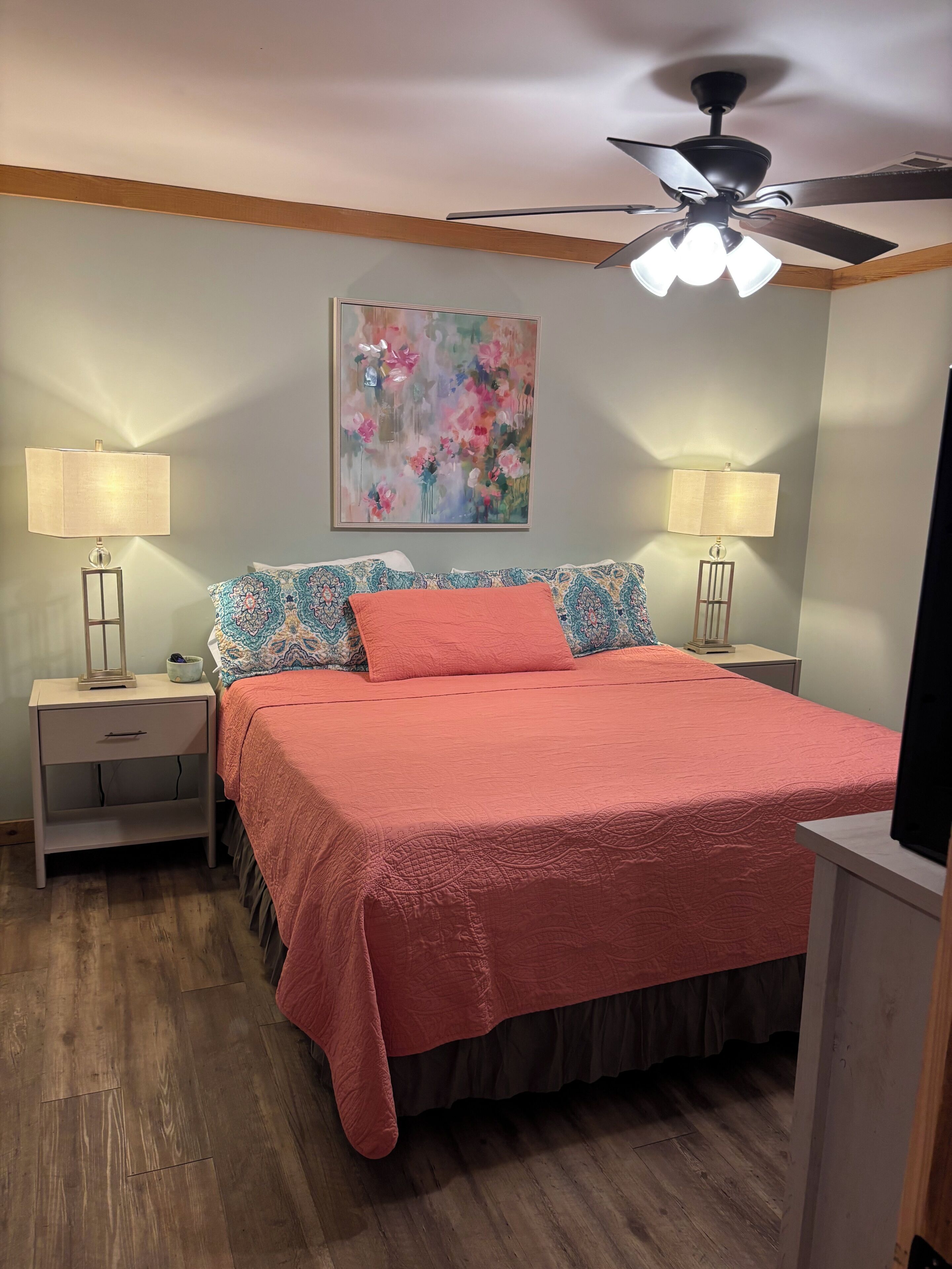 Une chambre à coucher avec un lit, des tables de chevet, des lampes, un ventilateur de plafond et un tableau floral accroché au mur.