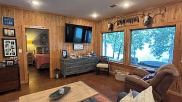 A cozy living room with a wooden interior, a flat-screen TV mounted on the wall, and a large window offering a view of trees.