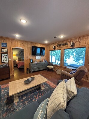 A cozy living room with a wooden interior, a flat-screen TV mounted on the wall, and a large window offering a view of trees.