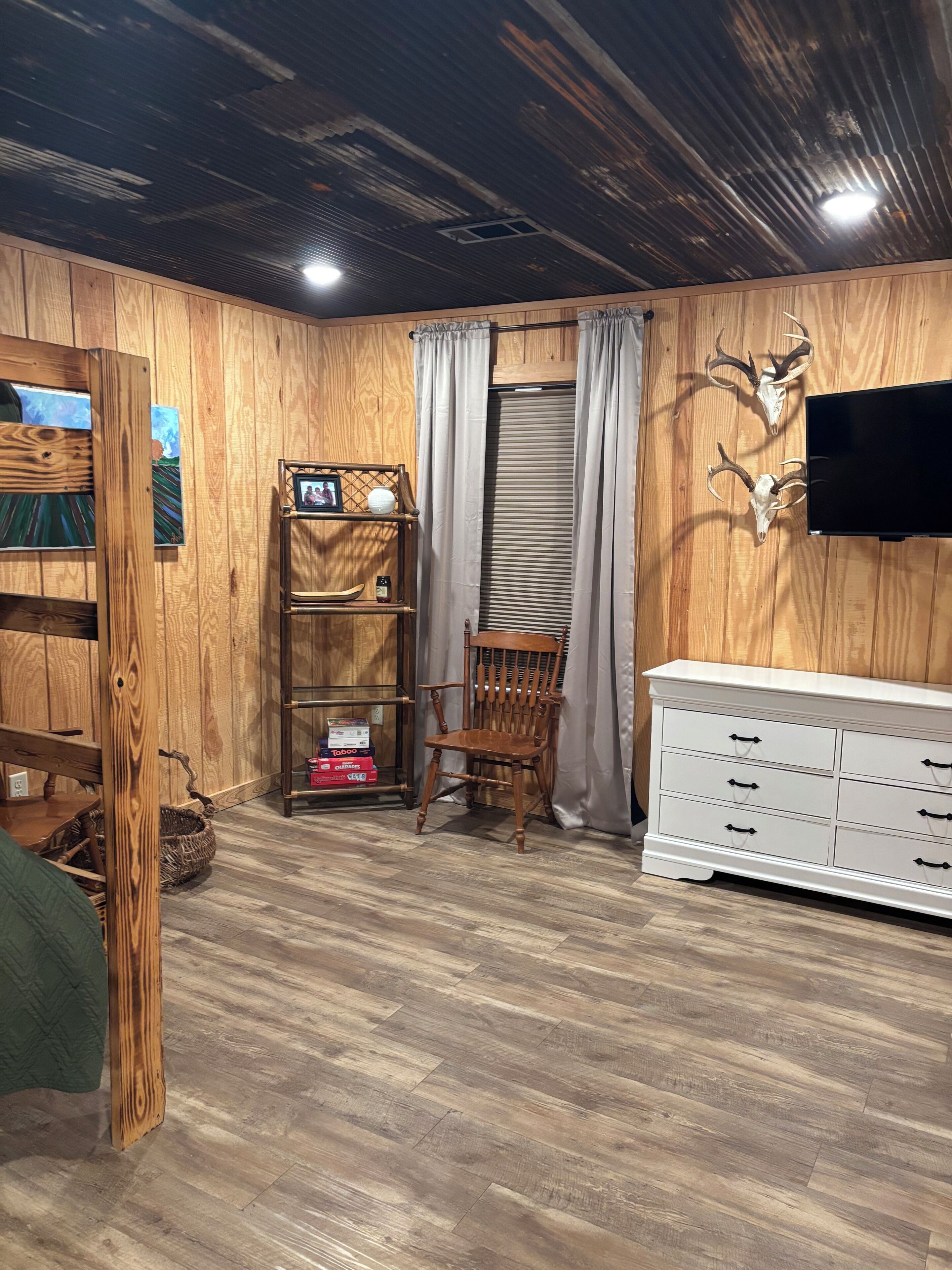 A room with wooden walls, a wooden floor, a white dresser, a wooden chair, a shelf with items, and a television mounted on the wall.