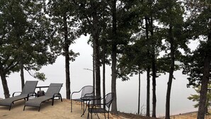 A serene outdoor seating area with lounge chairs and a table, surrounded by tall trees and overlooking a calm body of water.