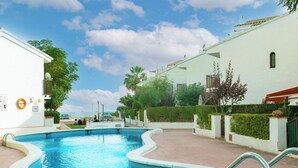Una piscina de borde curvo, rodeada por un camino empedrado y contigua a un edificio blanco con techo de tejas.