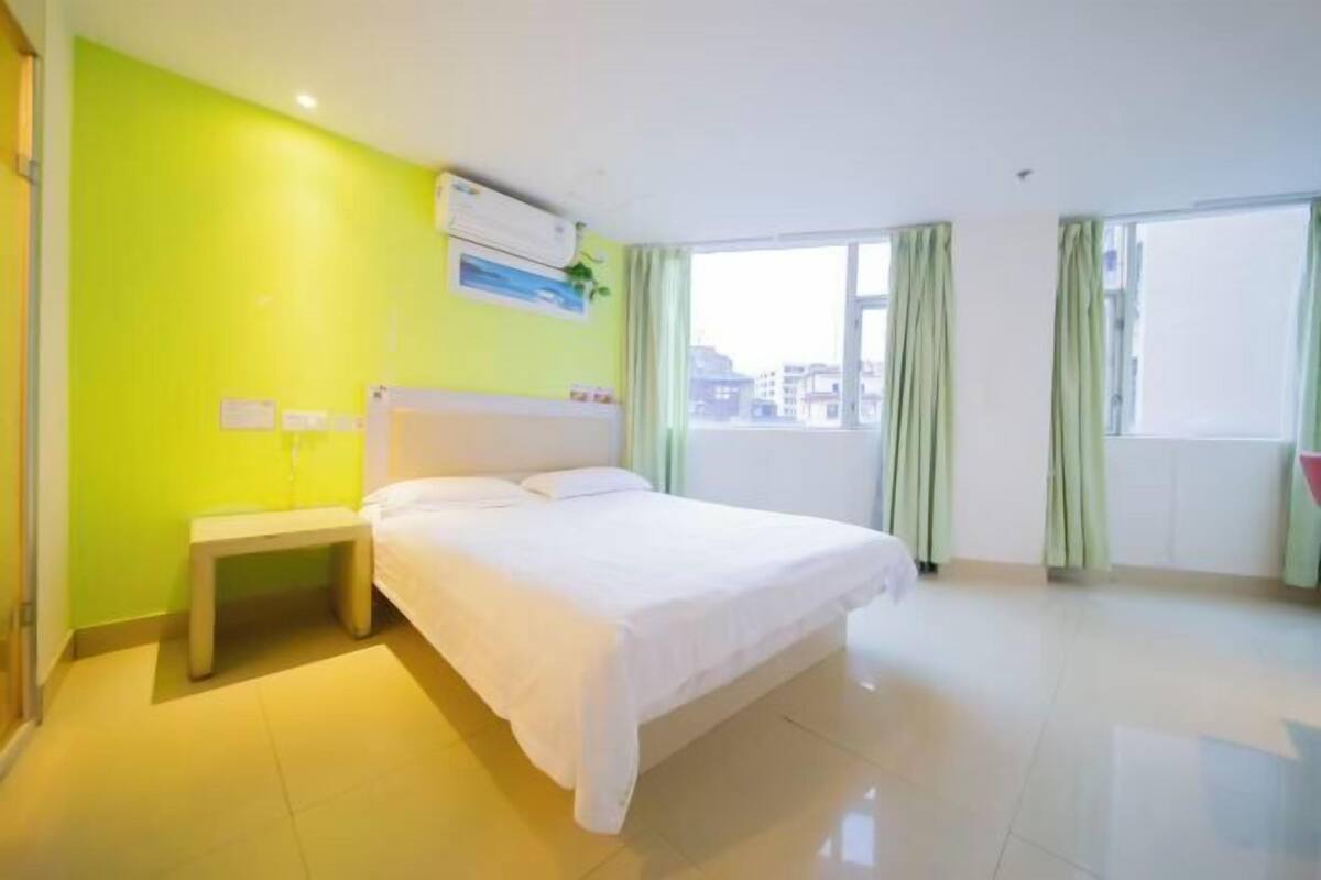 A hotel room with a bed, a bedside table, an air conditioning unit, and a window with curtains.