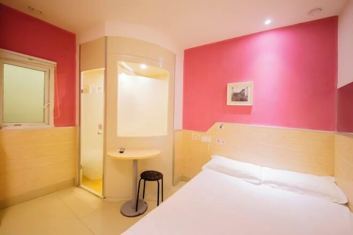 Una habitación de hotel con una cama, una mesita, un taburete, un espejo y un cuadro en la pared.
