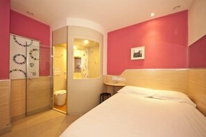 Un dormitorio con cama, una mesita, una silla, un armario y un baño visible a través de una puerta abierta.
