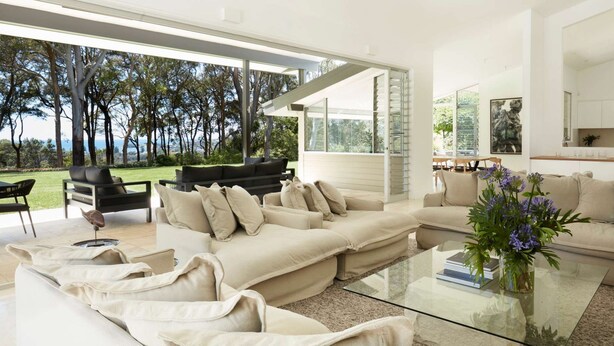 Un salon moderne avec une grande table en verre, des canapés de couleur beige et une vue sur un jardin et des arbres.