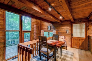 Interior de una cabaña de madera con una mesa de comedor, sillas, un tapete a cuadros y grandes ventanales que dan al bosque.