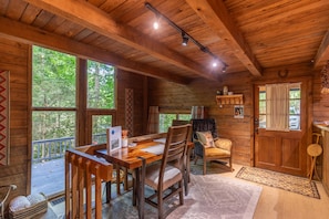 A wooden cabin interior with a dining table, chairs, and a view of the forest through large windows.