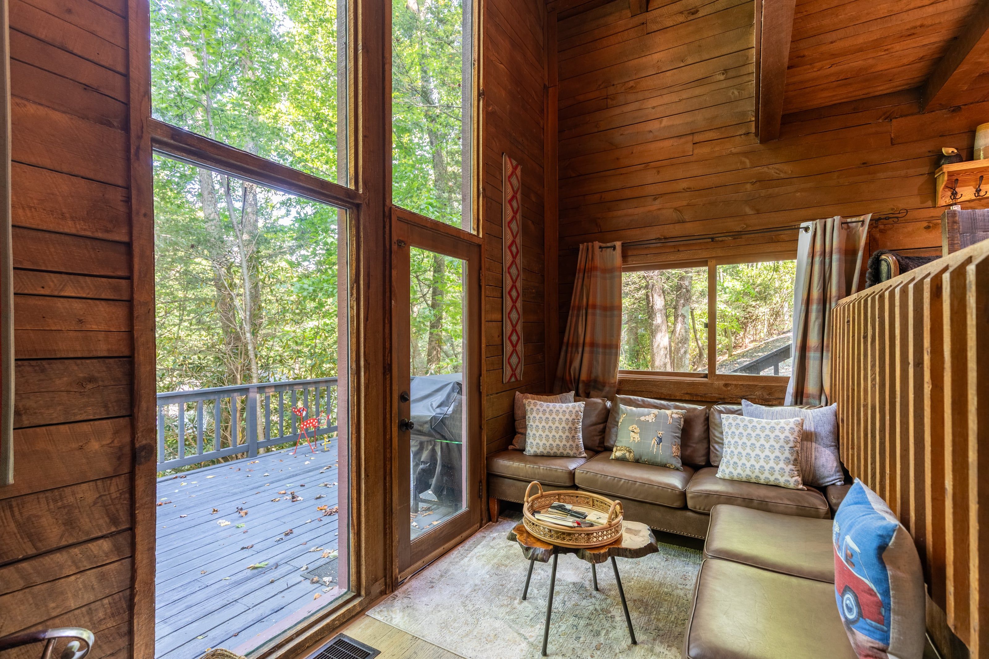A cozy cabin interior with a wooden floor, a sofa with patterned cushions, and a large window offering a view of the forest.